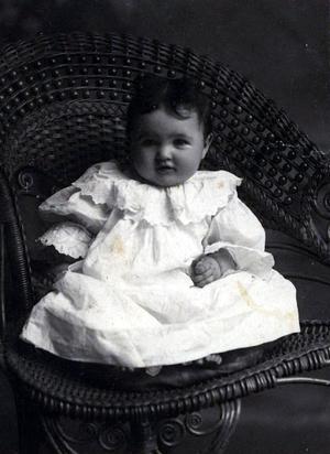 Baby in white dress posed on a wicker chair.