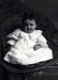 Photograph: Baby in white dress posed on a wicker chair.